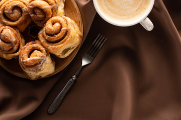 top view of fresh homemade cinnamon rolls on plate near coffee and fork on silk brown cloth