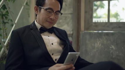 Asian groom sitting on a red chair using smartphone are smiling and happy in studio. Handheld shot. Real life - Powered by Adobe