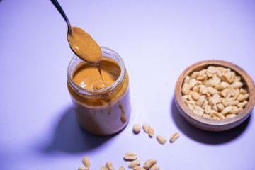 Spoon with peanut butter falling into a glass jar with some peanuts on purple background.