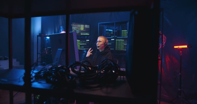 Caucasian woman in headset with mic talking while having call with client in dark room with monitors. Female worker of IT call-center in front of computer screen speaking and browsing online.