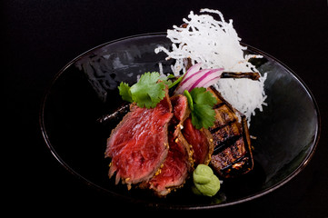 Grilled beef with eggplants wasabi and coriander in a black bowl, isolated on black backround