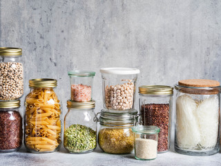 Glass jars with various cereals and seeds - peas split, sunflower and pumpkin seeds, beans, rice, pasta, oatmeal, lentils, bulgur on a grey background