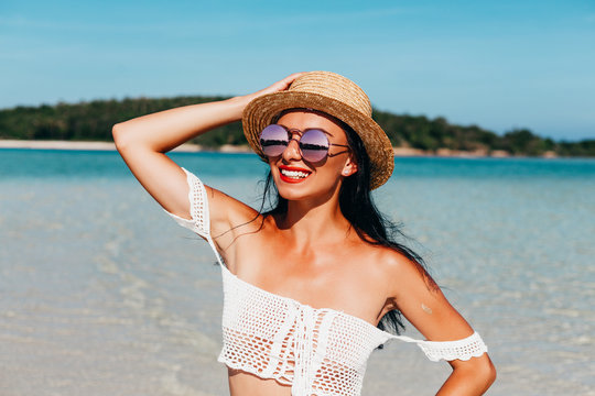 Summer time! Beautiful tanned lady in a handmade swimsuit, straw hat and glasses against the backdrop of a tropical island, smiles and enjoys the sun