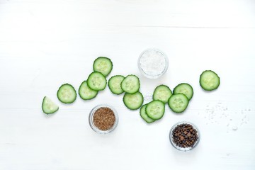 Sliced cucumber slices, salt, pepper, spices in glass bowls on a white wooden table. Freshness concept, vegetarianism, salad ingredients. Top view, flat lay, copy space for text.