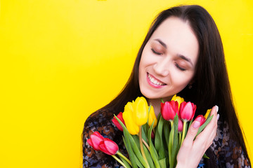 Portrait of a brunette woman with a bouquet of tulips on a yellow background. Birthday girl with flowers. Happy woman with a bouquet of flowers smiling closing her eyes.