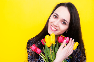 Obraz na płótnie Canvas A birthday girl with a bouquet of tulips looks out of the frame and smiles. Happy brunette with a bouquet of flowers. Florist with flowers on a yellow background.
