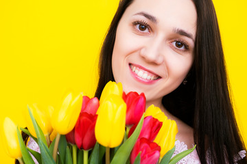 Woman portrait with a bouquet of flowers on a yellow background close-up. A brunette with tulips smiles and looks into the frame.