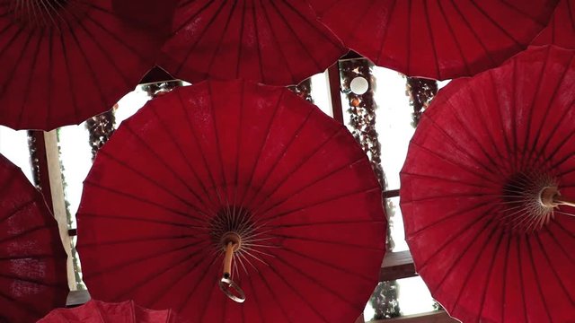 Traditional decorative umbrellas hanging under the roof of the building. A many red parasol is moving in wind under ceiling, Thailand.