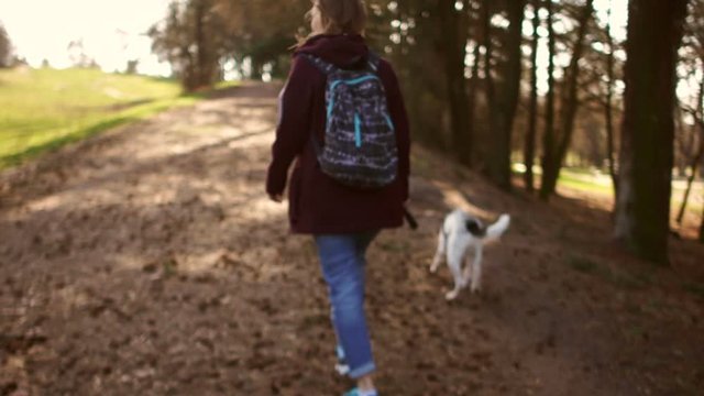 Young woman is walking with her dog on a leash in a spring city park. The girl is dressed in a burgundy jacket and wears a backpack on her back