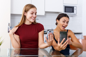 Two young girlfriends making selfie by phone and smiling at table