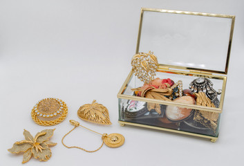 Open transparent glass box, inside which and next to it are gold and silver vintage jewelry with precious gem stones: brooches, pins, Swarovski crystals, chains, cameo etc