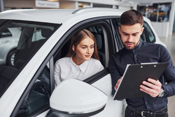 Fototapeta premium Woman testing car by help of male assistant that shows doments indoors in the salon
