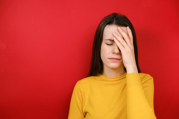 Frustrated woman holding hands on forehead being depressed regretting what she done having headache, wears yellow sweater, stands over red background. Negative human emotions and feelings concept