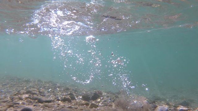 Slow motion underwater view of river bed. Rock fall into water causing bubbles and shock wave. Sun rays reflecting flowing particles under water surface