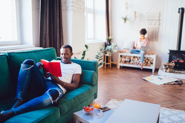 Black man reading book on sofa while woman on bench