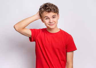 Portrait of pensive teen boy on grey background. Thoughtful teenager holding hand near head, looking at camera. Handsome caucasian funny child student in red t-shirt.