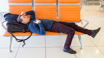 A 35-40 year old man sleeps on bench while waiting for his flight at the airport. A passenger is waiting for his plane at the airport.