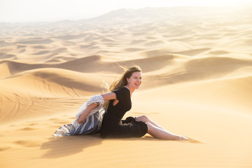 Fototapeta na wymiar Young beautiful woman sits tiredly on the hot sand of the desert under the hot rays of the eastern sun A girl in a black dress sits on the sand in the desert