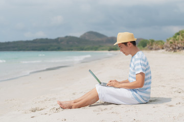 Asian man on holiday wearing a hat sit on the beach and working with a laptop.
