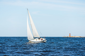 White sloop rigged yacht sailing on a clear day. Lighthouse in the background. Cloudy blue sky. Bay...