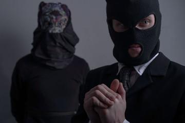 Close-up studio portrait, two robbers. A bandit in a black mask, and a jacket in the foreground looks to the side, another robber in a scary mask in the background. On a gray background.