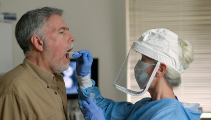 A mature Caucasian man in a clinical setting being swabbed by a healthcare worker in protective...