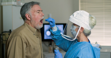 A mature Caucasian man in a clinical setting being swabbed by a healthcare worker in protective...
