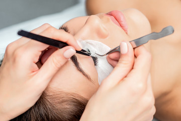 Cosmetologist glues artificial long eyelashes with tweezers on eye of woman while eyelash extension procedure.