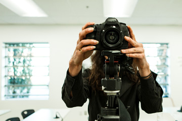 A close up and front view of a photographer using a DSLR camera mounted on a video tripod indoor. Behind lens shot of professional multimedia artist.