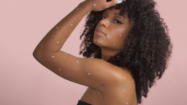 Mixed race black woman with curly hair covered by crystal makeup dancing on a pink background