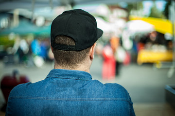 A large build man is seen from behind in selective focus, wearing blue denim shirt and black baseball cap at a street market with blurry stalls in background. - Powered by Adobe
