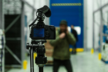 A close up selective focus shot of a professional DSLR camera and shotgun mic mounted on a tripod inside a warehouse, with blurry presenter in background.