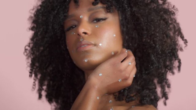 Mixed race black woman with curly hair covered by crystal makeup on pink background in studio touches her hair