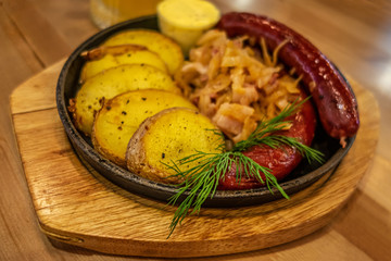 grill sausages garnished with baked potatoes and stewed sour cabbage on a cast iron pan on a table in a pub