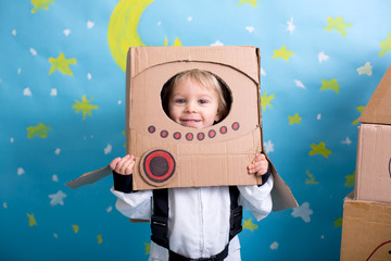 Sweet toddler boy, dressed as an astronaut, playing at home with cardboard rocket and handmade...