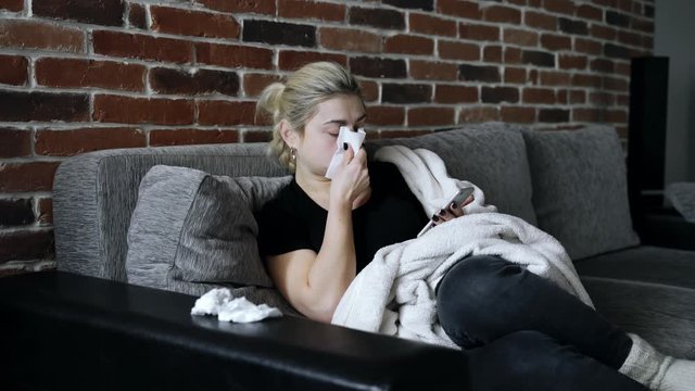 Health: A sick woman sits on a sofa at home and texting on smartphone. The girl  looks ill and sneezes and blows her nose and wipes her face with a napkin. She suffers from cough flu or coronavirus