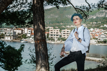 A girl with blue hair in a white jacket stands under the trees against the backdrop of a...