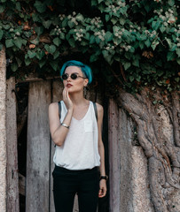 A woman in a white T-shirt and black jeans stands near a wooden gate at a concrete fence in greenery.