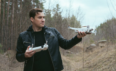 Young handsome man in casual clothes holding white drone and remote control in forest. Adult guy going to launch his drone in woodland.