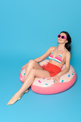 happy woman in sunglasses sunbathing while sitting on inflatable ring on blue background