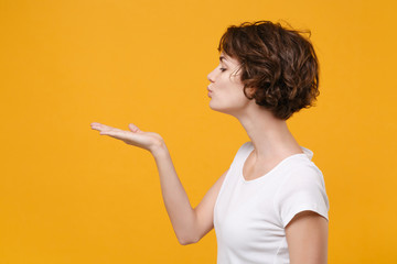 Side view of young brunette woman girl in white t-shirt posing isolated on yellow orange background studio portrait. People emotions lifestyle concept. Mock up copy space. Blowing sending air kiss.