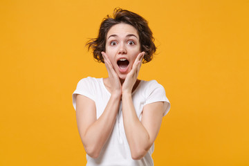 Shocked young brunette woman girl in white t-shirt posing isolated on yellow orange background studio portrait. People lifestyle concept. Mock up copy space. Keeping mouth open, put hands on cheeks.