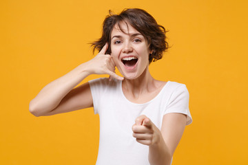Cheerful young brunette woman girl in white t-shirt isolated on yellow background. People lifestyle concept. Mock up copy space. Doing phone gesture says call me back, pointing index finger on camera.