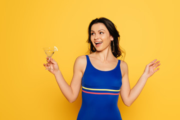 cheerful woman in blue swimsuit holding glass of cocktail and looking away isolated on yellow