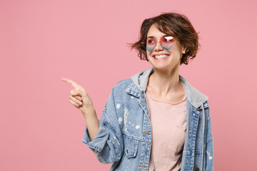 Smiling young brunette woman girl in casual denim jacket eyeglasses posing isolated on pastel pink background in studio. People lifestyle concept. Mock up copy space. Pointing index finger aside up.