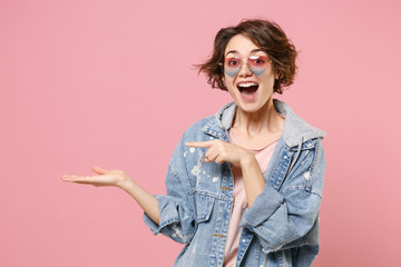 Excited young brunette woman girl in casual denim jacket eyeglasses posing isolated on pastel pink background in studio. People lifestyle concept. Mock up copy space. Pointing index finger hand aside.