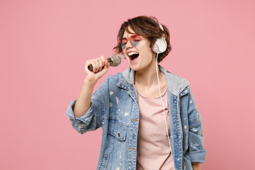 Pretty young brunette woman in casual denim jacket, eyeglasses isolated on pastel pink background. People lifestyle concept. Mock up copy space. Listen music with headphones sing song in microphone.