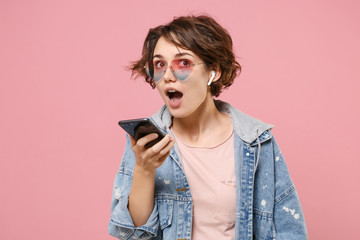 Young brunette woman in casual denim jacket, eyeglasses posing isolated on pastel pink background studio portrait. People lifestyle concept. Mock up copy space. Talking on mobile phone with air pods.