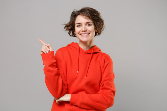 Smiling young brunette woman girl in casual red hoodie posing isolated on grey background studio portrait. People sincere emotions lifestyle concept. Mock up copy space. Point index finger aside up.