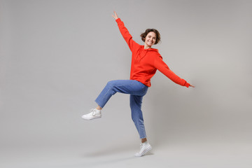 Fototapeta premium Side view of funny young brunette woman girl in casual red hoodie blue jeans posing isolated on grey wall background. People lifestyle concept. Mock up copy space. Dancing, spreading hands and legs.